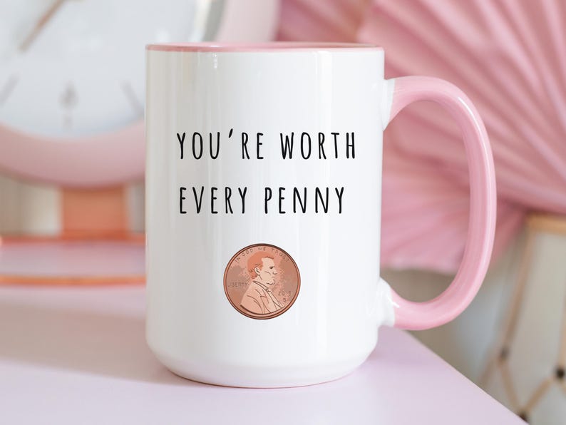 May include: White ceramic mug with a pink handle and interior. The mug features the text "YOU'RE WORTH EVERY PENNY" above a penny illustration. The mug is on a pink surface.