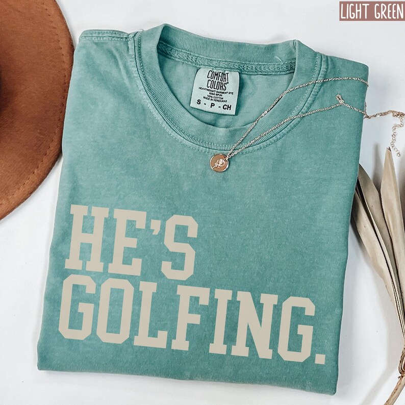 May include: A light green t-shirt with the text "HE'S GOLFING." printed in a cream color. The shirt is made of a soft, comfortable fabric and has a relaxed fit.