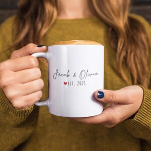 K&ouml;nnte beinhalten: Wei&szlig;e Keramik-Tasse mit Henkel, gef&uuml;llt mit einem Getr&auml;nk und mit Schaum bedeckt. Auf der Tasse stehen die Namen "Jacob & Olivia" in Schreibschrift, ein rotes Herz und "EST. 2021". Die Tasse wird von einer Person gehalten, die einen senffarbenen Pullover tr&auml;gt.