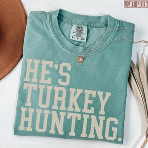 Puede incluir: Una camiseta verde claro con el texto "He's Turkey Hunting" en color crema. La camiseta est&aacute; hecha de un material suave y c&oacute;modo y tiene un corte holgado.