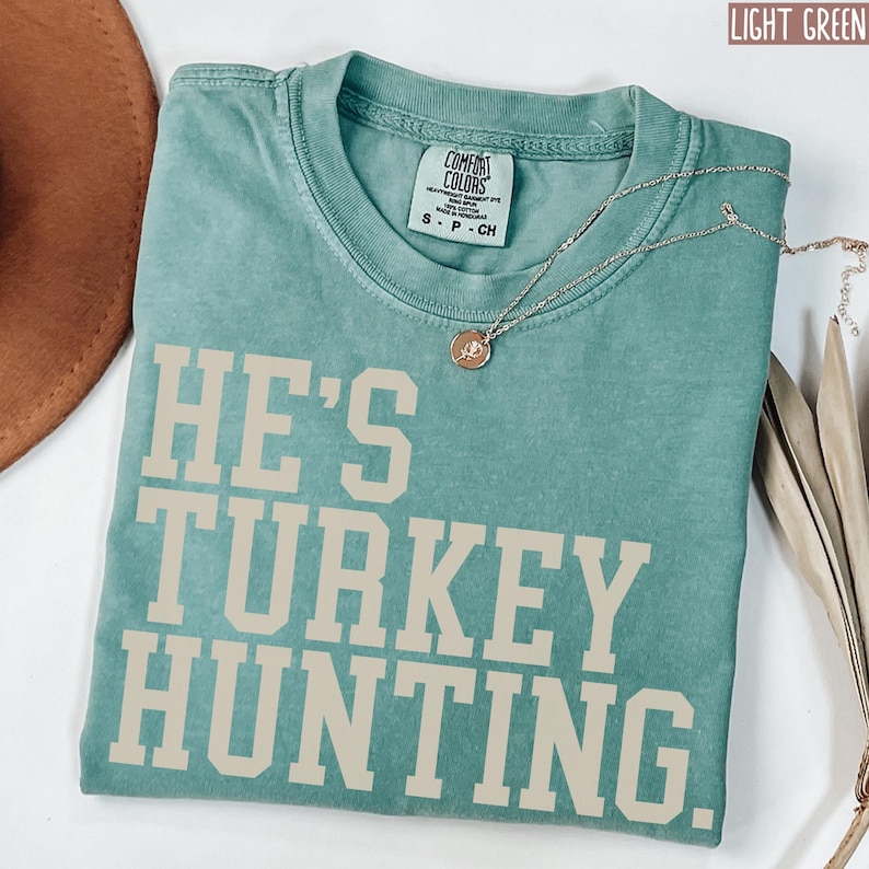 Puede incluir: Una camiseta verde claro con el texto "He's Turkey Hunting" en color crema. La camiseta est&aacute; hecha de un material suave y c&oacute;modo y tiene un corte holgado.