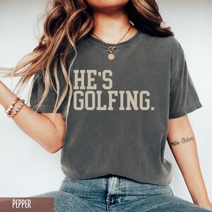 May include: A dark gray t-shirt with the text "HE'S GOLFING." printed in a light beige color.