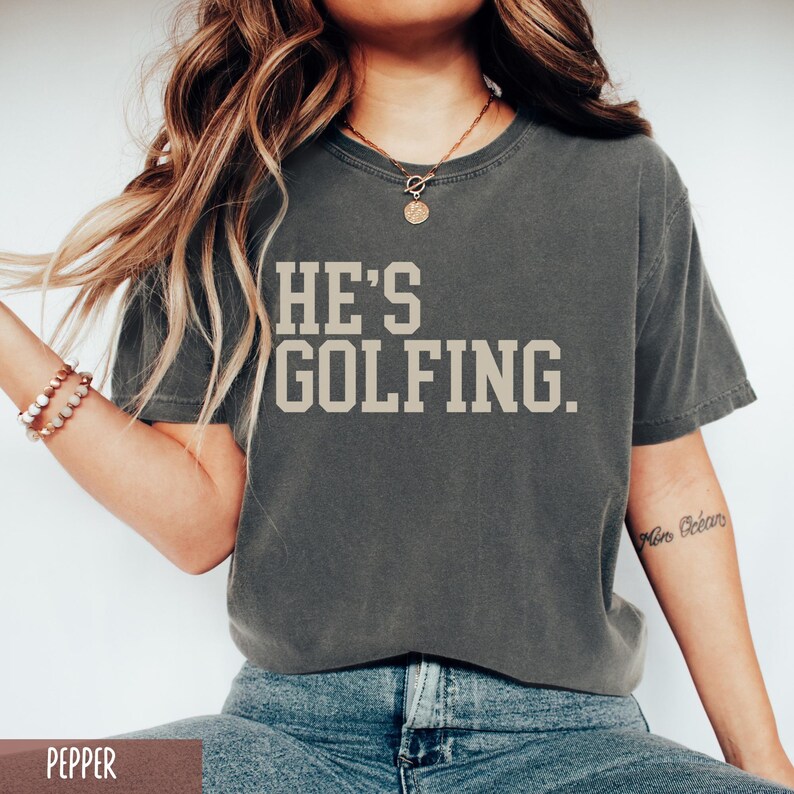 May include: A dark gray t-shirt with the text "HE'S GOLFING." printed in a light beige color.