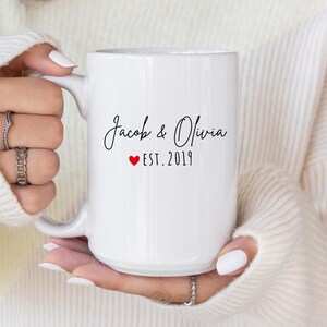 May include: White ceramic mug with the names "Jacob & Olivia" in a script font, a red heart, and "EST. 2019". The mug is held by a person wearing a white sweater.