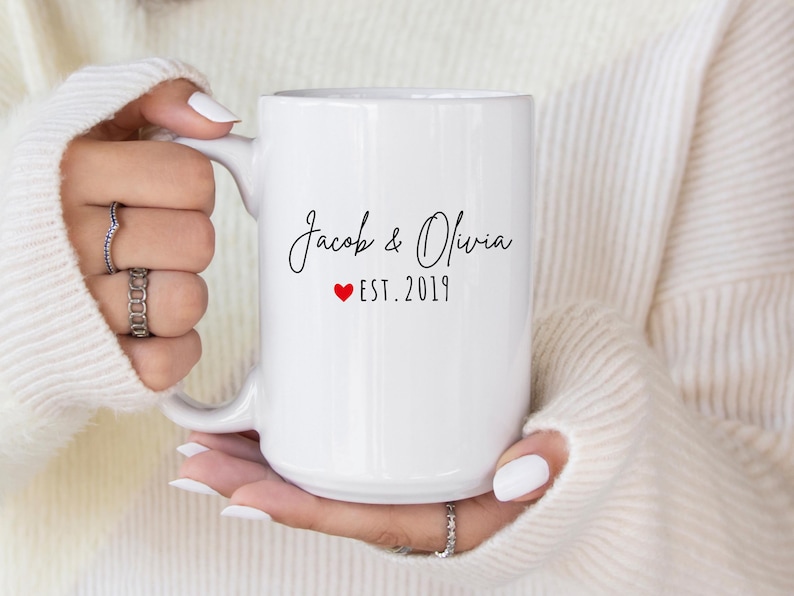 May include: White ceramic mug with the names "Jacob & Olivia" in a script font, a red heart, and "EST. 2019". The mug is held by a person wearing a white sweater.
