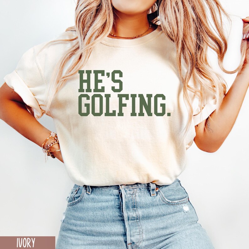 May include: A white t-shirt with the text "HE'S GOLFING." printed in green.