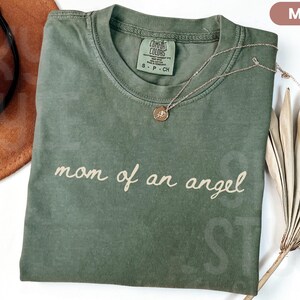 K&ouml;nnte beinhalten: Ein moosgr&uuml;nes T-Shirt mit dem Schriftzug "mom of an angel" in Schreibschrift. Eine goldfarbene Halskette mit Anh&auml;nger liegt auf dem Shirt. Ein brauner Hut und getrocknete Palmbl&auml;tter befinden sich im Hintergrund.