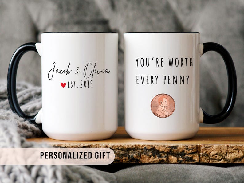 May include: Two white ceramic mugs with black handles and rims. One mug has the names "Jacob & Olivia" with a red heart and "EST. 2019". The other mug says "YOU'RE WORTH EVERY PENNY" with a penny illustration. "PERSONALIZED GIFT" is written at the bottom.