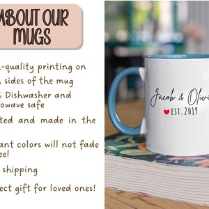 May include: White ceramic mug with a blue handle and interior. The mug features the names "Jacob & Olivia" with a red heart and "EST. 2019" printed on it. The mug is sitting on a stack of books.