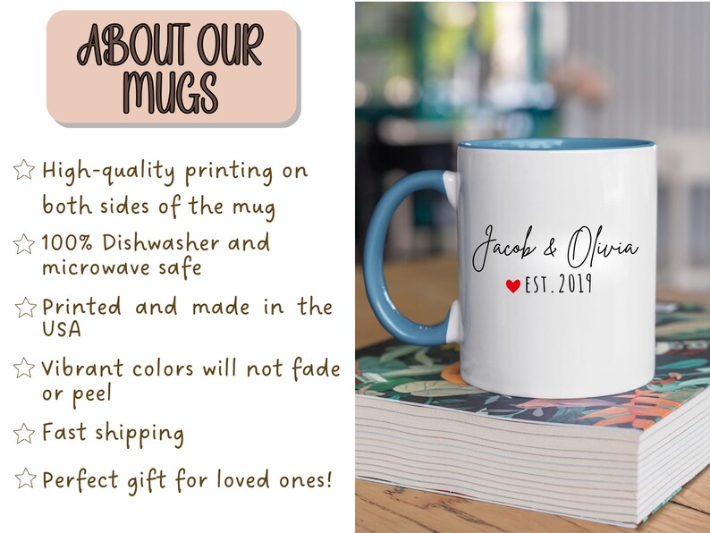 May include: White ceramic mug with a blue handle and interior. The mug features the names "Jacob & Olivia" with a red heart and "EST. 2019" printed on it. The mug is sitting on a stack of books.