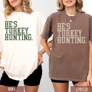 Puede incluir: Dos camisetas con el texto "He's Turkey Hunting." La camiseta de la izquierda es de color marfil y la camiseta de la derecha es de color espresso.