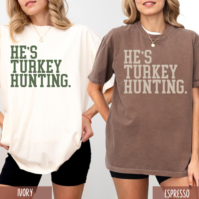 Puede incluir: Dos camisetas con el texto "He's Turkey Hunting." La camiseta de la izquierda es de color marfil y la camiseta de la derecha es de color espresso.