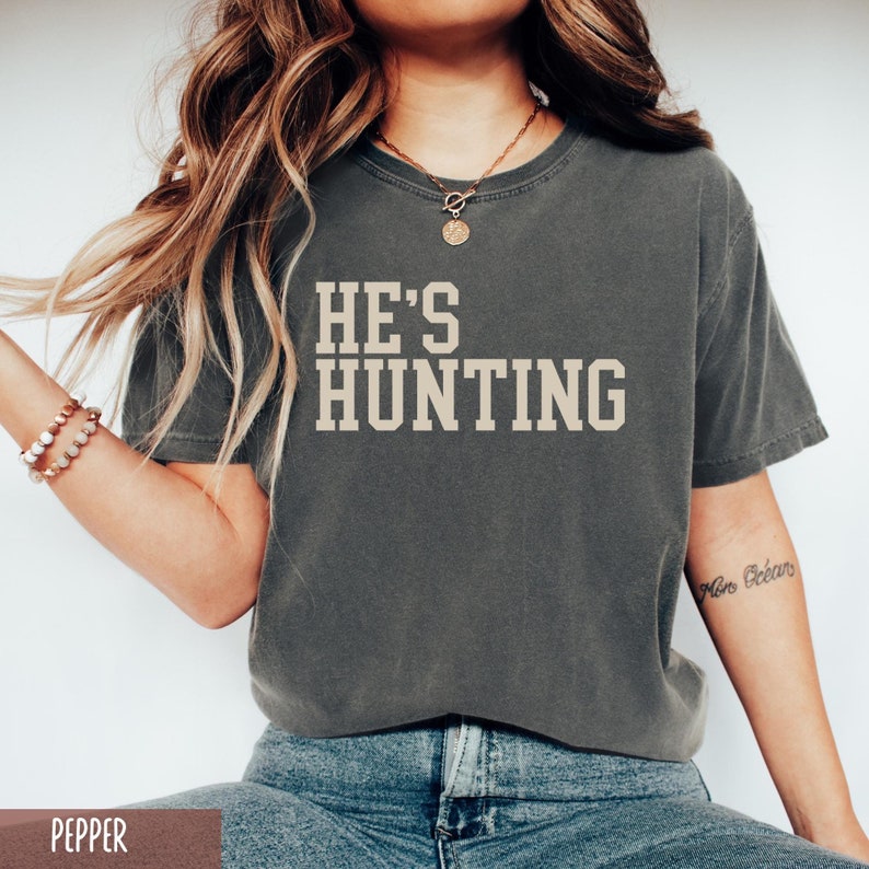 May include: A gray t-shirt with the text "HE'S HUNTING" printed in white on the front. The shirt is being worn by a person with long brown hair.