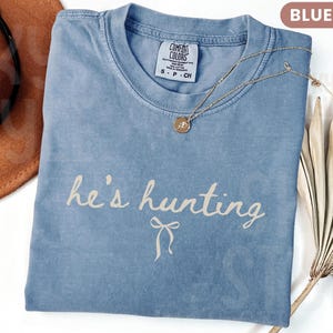 以下が含まれることがあります： 「he's hunting」という筆記体の文字が入った、ブルーデニムのComfort Colors Tシャツ。ペンダント付きのゴールドネックレスがシャツの上にあります。茶色の帽子が左上に写っています。
