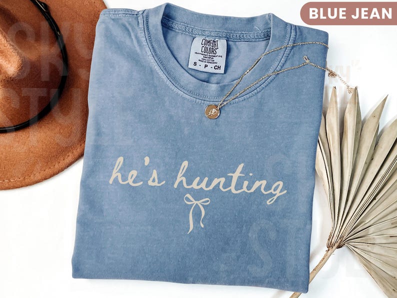 以下が含まれることがあります： 「he's hunting」という筆記体の文字が入った、ブルーデニムのComfort Colors Tシャツ。ペンダント付きのゴールドネックレスがシャツの上にあります。茶色の帽子が左上に写っています。