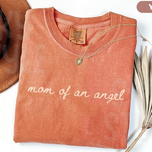 K&ouml;nnte beinhalten: Ein yamfarbenes T-Shirt mit dem Schriftzug "mom of an angel" in Schreibschrift. Eine goldene Halskette mit Anh&auml;nger liegt auf dem Shirt. Ein brauner Hut und getrocknete Palmbl&auml;tter sind ebenfalls im Bild.