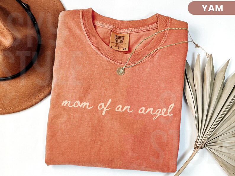 K&ouml;nnte beinhalten: Ein yamfarbenes T-Shirt mit dem Schriftzug "mom of an angel" in Schreibschrift. Eine goldene Halskette mit Anh&auml;nger liegt auf dem Shirt. Ein brauner Hut und getrocknete Palmbl&auml;tter sind ebenfalls im Bild.