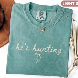 以下が含まれることがあります： 「he's hunting」という筆記体の文字が入った、明るい緑色のTシャツ。丸いペンダントが付いたシルバーのネックレスがシャツの上にあります。左上には茶色の帽子が見えます。シャツは柔らかい素材で作られています。