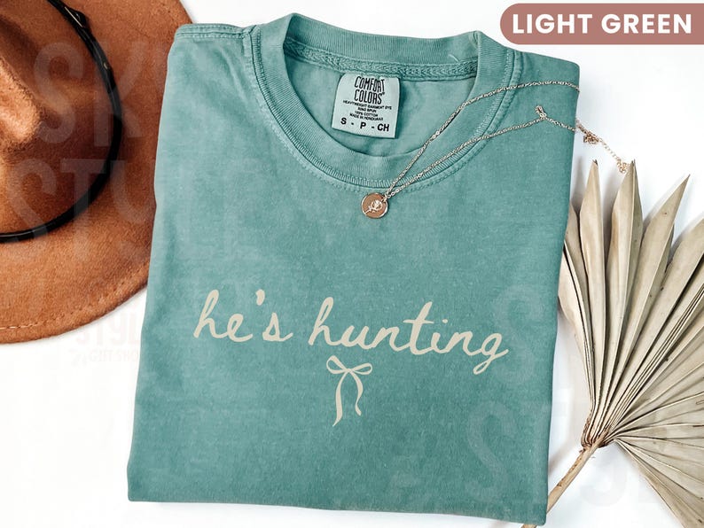 以下が含まれることがあります： 「he's hunting」という筆記体の文字が入った、明るい緑色のTシャツ。丸いペンダントが付いたシルバーのネックレスがシャツの上にあります。左上には茶色の帽子が見えます。シャツは柔らかい素材で作られています。
