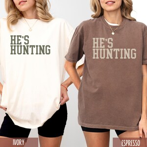 May include: Two oversized t-shirts with the text "HE'S HUNTING" in a green and white font. The shirts are shown on models wearing black shorts. The shirt on the left is ivory and the shirt on the right is espresso.