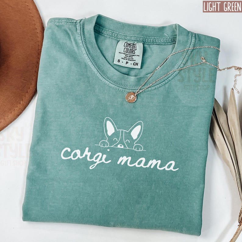 May include: Light green t-shirt with a white outline of a corgi peeking over the top and the text "corgi mama" underneath. The shirt is made of a soft, comfortable material.