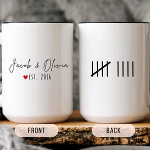 May include: White ceramic mug with a black handle and rim. The front of the mug has the names "Jacob & Olivia" with a red heart and "EST. 2016". The back has a tally mark design. The mug is on a wooden surface.