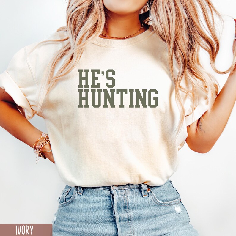 May include: A white t-shirt with the text "HE'S HUNTING" printed in green on the front. The shirt is being worn by a person with long blonde hair. The shirt is ivory in color.
