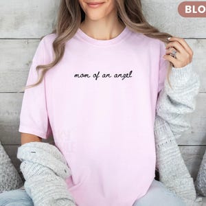 K&ouml;nnte beinhalten: Hellrosa T-Shirt mit dem schwarzen Schriftzug "mom of an angel". Das Shirt hat einen l&auml;ssigen Stil. Das Wort "BLOSSOM" befindet sich in einem kleinen Rechteck oben rechts.