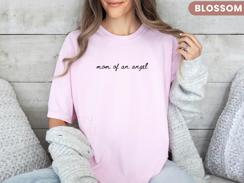 K&ouml;nnte beinhalten: Hellrosa T-Shirt mit dem schwarzen Schriftzug "mom of an angel". Das Shirt hat einen l&auml;ssigen Stil. Das Wort "BLOSSOM" befindet sich in einem kleinen Rechteck oben rechts.