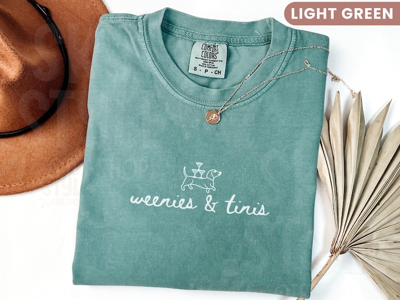 Może przedstawiać: Jasnozielona koszulka Comfort Colors z napisem "weenies & tiris" i grafiką psa. Złoty naszyjnik z wisiorkiem spoczywa na koszulce. Brązowy kapelusz i suszone liście znajdują się w tle.