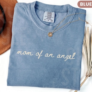 K&ouml;nnte beinhalten: Blaues Comfort Colors T-Shirt in Jeansoptik mit dem Schriftzug "mom of an angel" in Schreibschrift. Eine goldene Halskette mit Anh&auml;nger liegt auf dem Shirt. Ein brauner Hut ist im Hintergrund zu sehen.