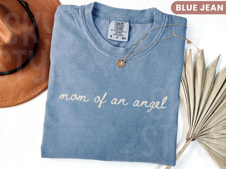 K&ouml;nnte beinhalten: Blaues Comfort Colors T-Shirt in Jeansoptik mit dem Schriftzug "mom of an angel" in Schreibschrift. Eine goldene Halskette mit Anh&auml;nger liegt auf dem Shirt. Ein brauner Hut ist im Hintergrund zu sehen.
