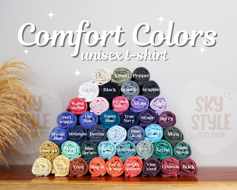 May include: A variety of colors of t-shirts rolled up in a pyramid shape. The colors include white, black, khaki, blue, green, red, orange, yellow, and purple. The text "Comfort Colors unisex t-shirt" is at the top of the image. The text "Sky Style Gift Shop" is at the bottom right of the image.