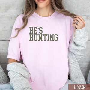 May include: A pink t-shirt with the text "HE'S HUNTING" in dark green letters.
