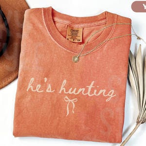 以下が含まれることがあります： 「he's hunting」という筆記体のフレーズが入った、ヤム色のTシャツ。ペンダント付きのゴールドネックレスがシャツの上にあります。茶色の帽子と乾燥したヤシの葉も画像にあります。