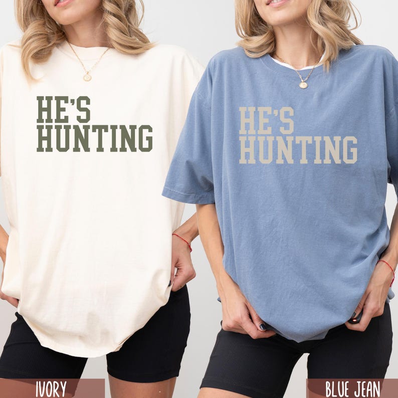 May include: Two oversized t-shirts with the text "He's Hunting" in a vintage style font. The shirt on the left is ivory and the shirt on the right is blue jean.