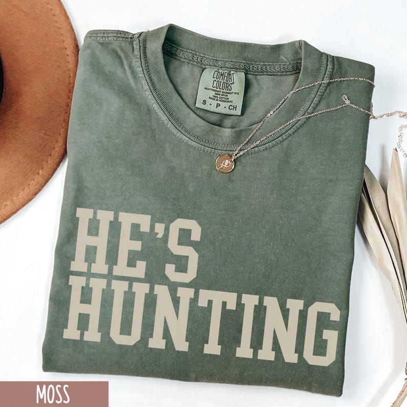 May include: A moss green t-shirt with the text "HE'S HUNTING" in a cream color. The shirt is made of a soft, comfortable fabric and has a relaxed fit.