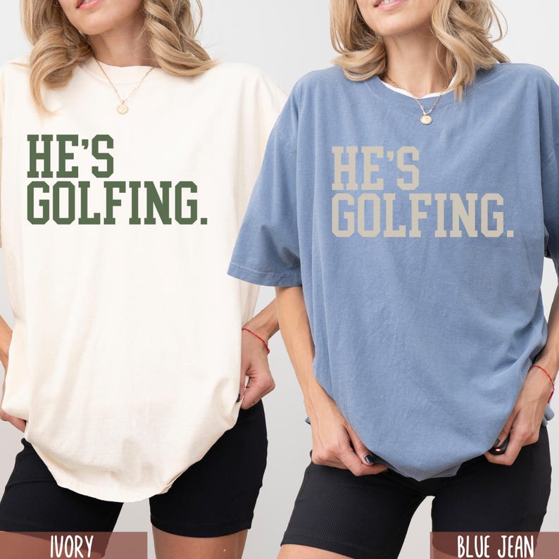 May include: Two oversized t-shirts with the text "He's golfing." in different colors. The shirt on the left is ivory with green text. The shirt on the right is blue jean with beige text.