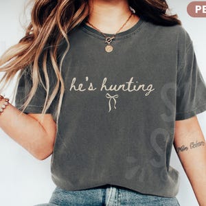 以下が含まれることがあります： 「he's hunting」の筆記体とリボンが描かれたペッパーカラーのTシャツ。 ゆったりとしたフィット感で、ヴィンテージ感のあるデザインです。 モデルは青いジーンズと合わせています。