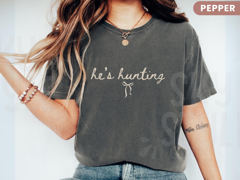 以下が含まれることがあります： 「he's hunting」の筆記体とリボンが描かれたペッパーカラーのTシャツ。 ゆったりとしたフィット感で、ヴィンテージ感のあるデザインです。 モデルは青いジーンズと合わせています。
