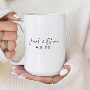 K&ouml;nnte beinhalten: Wei&szlig;er Keramikbecher mit den Namen "Jacob & Olivia" in Schreibschrift, einem roten Herz und "EST. 2021". Der Becher wird von einer Person gehalten, die einen wei&szlig;en Pullover und Ringe tr&auml;gt.
