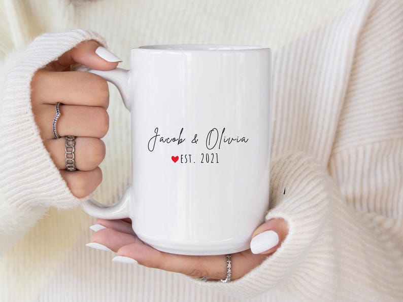 K&ouml;nnte beinhalten: Wei&szlig;er Keramikbecher mit den Namen "Jacob & Olivia" in Schreibschrift, einem roten Herz und "EST. 2021". Der Becher wird von einer Person gehalten, die einen wei&szlig;en Pullover und Ringe tr&auml;gt.