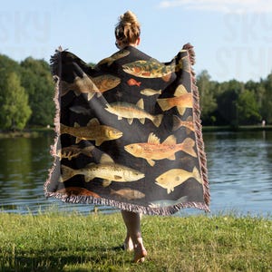 May include: A black woven throw blanket with a pattern of colorful fish. The blanket has fringed edges and is being held by a person.