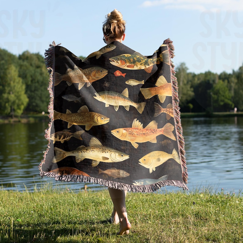 May include: A black woven throw blanket with a pattern of colorful fish. The blanket has fringed edges and is being held by a person.