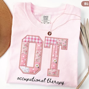 May include: A light pink t-shirt with the letters "OT" appliqued on the front. The letters are made of floral, polka dot, and gingham fabrics. The words "occupational therapy" are written in script below the letters. A gold necklace is draped across the shirt.