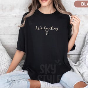 以下が含まれることがあります： 黒いTシャツ。筆記体で「he's hunting」の文字とリボンのグラフィックがプリントされています。Tシャツは無地の黒色で、半袖です。テキストは薄いベージュ色です。
