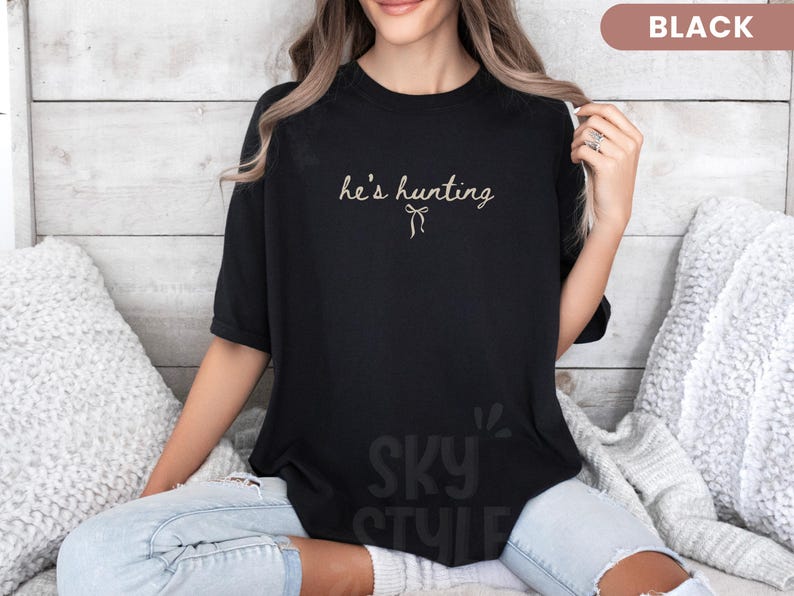 以下が含まれることがあります： 黒いTシャツ。筆記体で「he's hunting」の文字とリボンのグラフィックがプリントされています。Tシャツは無地の黒色で、半袖です。テキストは薄いベージュ色です。