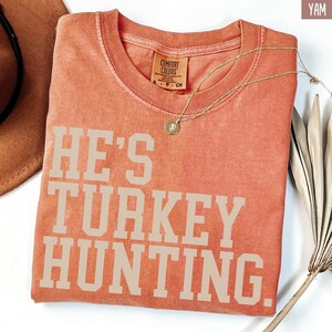 Puede incluir: Una camiseta naranja quemada con el texto "HE'S TURKEY HUNTING." impreso en blanco.