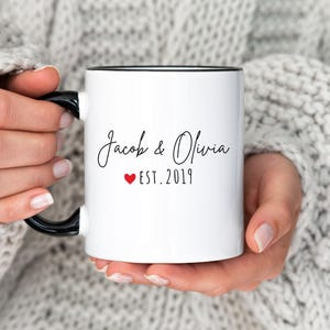 May include: White ceramic mug with black handle and rim, featuring the names "Jacob & Olivia" in elegant script, a red heart, and the year "EST. 2019". The mug is held by a person wearing a gray sweater.