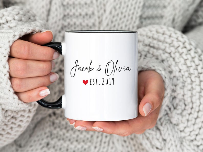 May include: White ceramic mug with black handle and rim, featuring the names "Jacob & Olivia" in elegant script, a red heart, and the year "EST. 2019". The mug is held by a person wearing a gray sweater.